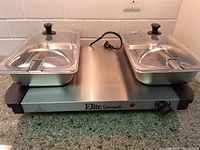 Front view showing the Elite Gourmet electric plate warmer with two stainless steel serving dishes topped with clear plastic covers having black knob handles.