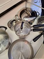 Photo showing a set of five cookware items on a countertop: two large soup pots with glass lids, one medium pot with a metal steamer insert and glass lid, one smaller pot with lid, and one frying pan with a black handle.