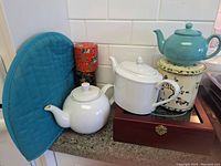 Three teapots arranged on and around a wooden tea box with three metal tea tins and a blue quilted teapot cozy beside them on countertop with white tile backsplash.