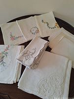 Assortment of white and off-white embroidered napkins and tablecloths folded on a dark wood surface showing floral embroidery and lace edges.