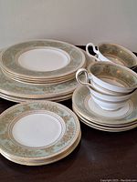 Set of four dinner plates, lunch plates, four cups and saucers with sage green and gold Wedgwood Gold Columbia pattern arranged on a table, showing the detailed gold decorative border and matching set pieces.