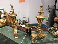 Front and side view of pair of brass andirons on green countertop with surrounding items
