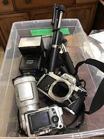 Top view photo showing Nikon FE camera body, Sony digital camera, Sunpak flash unit, and two selfie sticks inside a plastic container.