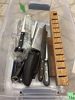 Top view of plastic lidded bin containing various knives, wooden knife block, and black handle sharpener.