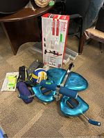 Photo of aerobic step in box, Total Tiger exercise handles, volleyball, and other accessories on the floor next to chair.