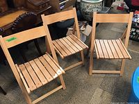 Three wood veneer folding chairs set up side by side, showing front and side views of slatted seats and solid backrests.