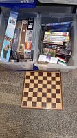 Two large plastic bins containing various puzzles, boxed checkers and chess sets; wooden chessboard placed in front of bins.
