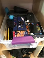 Plastic storage box with various gaming accessories including a Space Invaders mini arcade machine, gaming controllers, and cables.