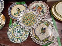 Five hand painted European plates shown with varied decorative motifs: farmer figure, floral design, geometric pattern, and two rooster designs. Plates show chips and crazing.