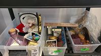 Plastic bins with assorted jewelry boxes and decorative items including porcelain doll figurine, blue decorative bottle, and several small boxes placed on a shelf.