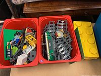 Two red bins filled with assorted gray track pieces, mixed system bricks and a green baseplate, plus a green/white train locomotive