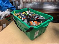 Green plastic bin filled with assorted LEGO pieces, showing a variety of bricks, wheels, and vehicle parts in mixed colors.