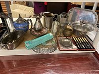 Image showing all metal ware dishes and items including teapot, pitchers, trays, coasters, and boxed cutlery on a shelf.
