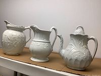 Full view of all three white ceramic pitchers on a cardboard surface, showing relative sizes and shapes