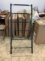 Front-facing view of the black metal plant stand with five levels, showing its form and size in relation to boxed items behind.