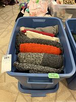 Blue lidded storage bin filled with folded sweaters and jackets in multiple colors and textures.