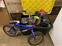 Photo showing blue children's bicycle next to a black storage bin containing two remote controlled trucks with large tires and a yellow lid.