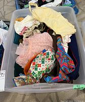 Clear lidded plastic bin filled with assorted vintage baby and children's clothing items including tops, dresses, and rompers in various colors and patterns, showing signs of wear and stains.