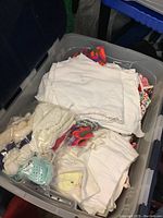 Top view of plastic tote showing mostly white folded cloth diapers or coverlets mixed with some colorful patterned fabrics and knitted hats inside the bin.