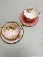 Two vintage porcelain teacup and saucer sets, one pink with gold floral details and one solid textured pink with floral interior, displayed on table.