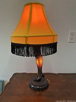 Photo of illuminated leg lamp showing yellow lampshade with black fringes, leg base with fishnet stocking, and high heel shoe.