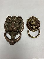 Front view of both brass lion head pieces: door knocker and drawer pull side by side on a flat surface.