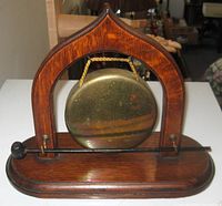 Front view of wood framed brass gong with black metal striker on wooden base, showing intact brass gong and polished wood.