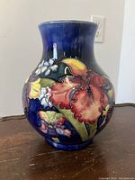 Front angle showing swollen orchid floral design in red, yellow, white on deep blue background, with bulbous shape narrowing at neck and flared rim.