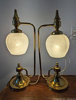 Pair of brass Hollywood Regency table lamps with opaque cylindrical white art glass shades, both shown illuminated on a wooden surface.