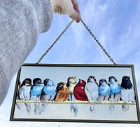 Photo showing the painted glass with colorful birds sitting on a branch, held by the hanging chain outside against the sky.