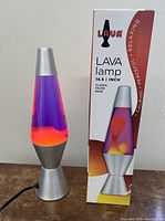 Front angled view of the vintage lava lamp next to its original boxed packaging, showing purple liquid and red lava shapes inside silver base.