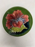 Top view showing circular dish with red hibiscus flower and green leaf design on dark green background.