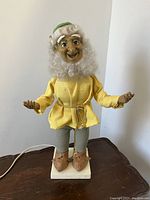 Full front view of vintage mechanical gnome figurine standing on table, showing yellow tunic, striped pants, green hat, white hair and base with cord.
