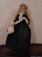 Full view of the porcelain doll in a dark dress with white fur trim, standing on a surface against a wall.