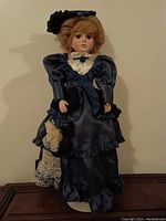 Front view of porcelain collectible doll wearing navy blue satin Victorian-style dress with lace details and bonnet, on stand.