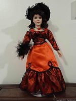 Porcelain collectible doll standing on a wooden surface wearing an orange satin dress with black lace and flower accents. The doll has dark curly hair and a black lace hat decorated with orange flowers. Visible details include feather trim on sleeve and painted facial features.