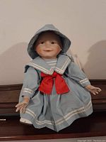 Front view of porcelain collectible doll dressed in light blue sailor dress with white trim and a large red bow, seated on a wooden surface.