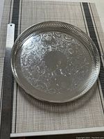 Full view of round silver tray with pierced border and intricate floral embossed design, placed next to ruler for scale.