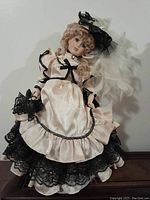 Front and angled view of the porcelain collectible doll showing the Victorian-style pink satin dress with black lace trim and curly blonde hair, black feathered hat.
