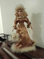 Front view of porcelain doll with curly blonde hair, brown satin dress trimmed with off-white fur, holding a matching fur muff.