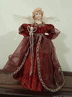 Front view of porcelain angel doll wearing red dress with sheer overlay and holding a beaded scepter, standing on wooden surface.