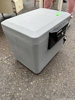 Closed gray plastic locking box on ground showing textured surface, handle on top, black lock with key inserted on front.
