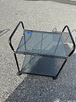 Front angle view of the black metal rolling shelf showing two perforated mesh shelves, tubular frame, and castor wheels.