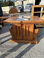 Front view of solid oak magazine table with glass inset top and carved magazine slots on the sides, outdoors on pavement with other furniture in background.