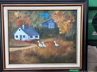 Framed painting depicting a white house with a grey roof and chimney, a darker building behind, autumn trees and three white geese, in a gold and black frame.