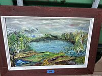Framed oil painting showcasing a natural landscape with a lake, greenery, and cloudy sky
