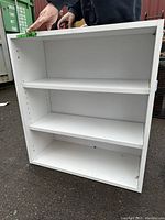 Front view of the white particle board shelving unit showing two adjustable shelves inside and visible peg holes for shelf height adjustments.