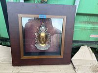 Wooden square frame with a glass front containing a decorative golden Buddha head with silver adornments on a reddish fabric background placed outdoors on cardboard.