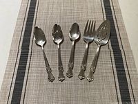 Five silver serving utensils laid on a table runner showing three spoons, a fork, and a pierced server with decorative floral patterns on handles and utensil heads.