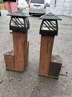 Pair of yard lights photographed outside on pavement, showing black metal tops with wave-like designs and wooden block bases with visible weathering.